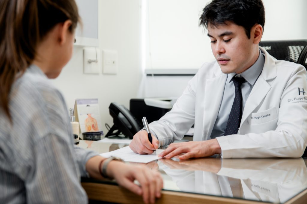 Oncologista Dr. Hugo Tanaka em consulta com uma paciente, fazendo anotações sobre o tratamento oncológico e explicando sobre a perda de peso no tratamento do câncer.