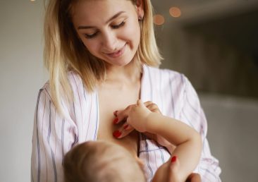 Back view of adorable six month old infant drinking breast milk. Attractive young European woman in home clothing cradling her baby daughter in arms, breastfeeding her, enjoying deep connection