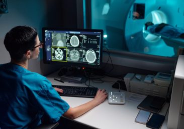 Medical professional reviewing patient's MRI images on computer monitor with patient in MRI machine in background. Hospital equipment and technology setup visible