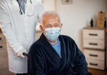 Happy mature man in wheelchair wearing protective face mask at nursing home. His doctor is standing behind him.