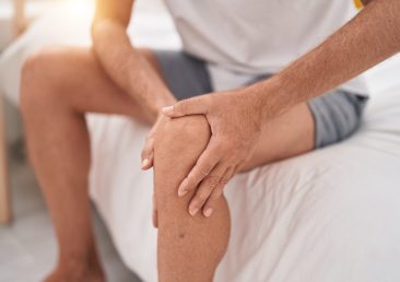 Young blond man suffering for knee injury sitting on bed at bedroom